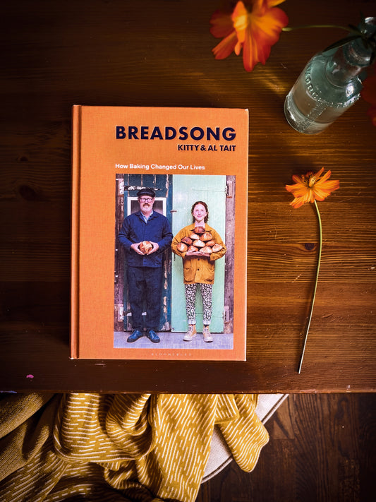 Orange Cosmos flowers happliy surrounding the book Breadsong, Cover is a warm orange with photo of Kitty and Al Tait holding bread