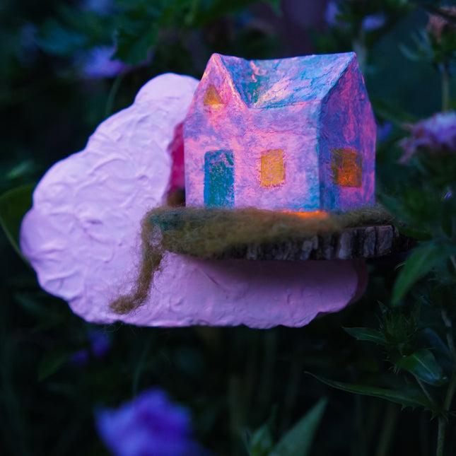 Small house sculpture on a cloud with glowing windows, surrounded by purple flowers.
